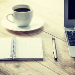 a cup of black coffee on a sourcer next to anote book and a pen and laptop